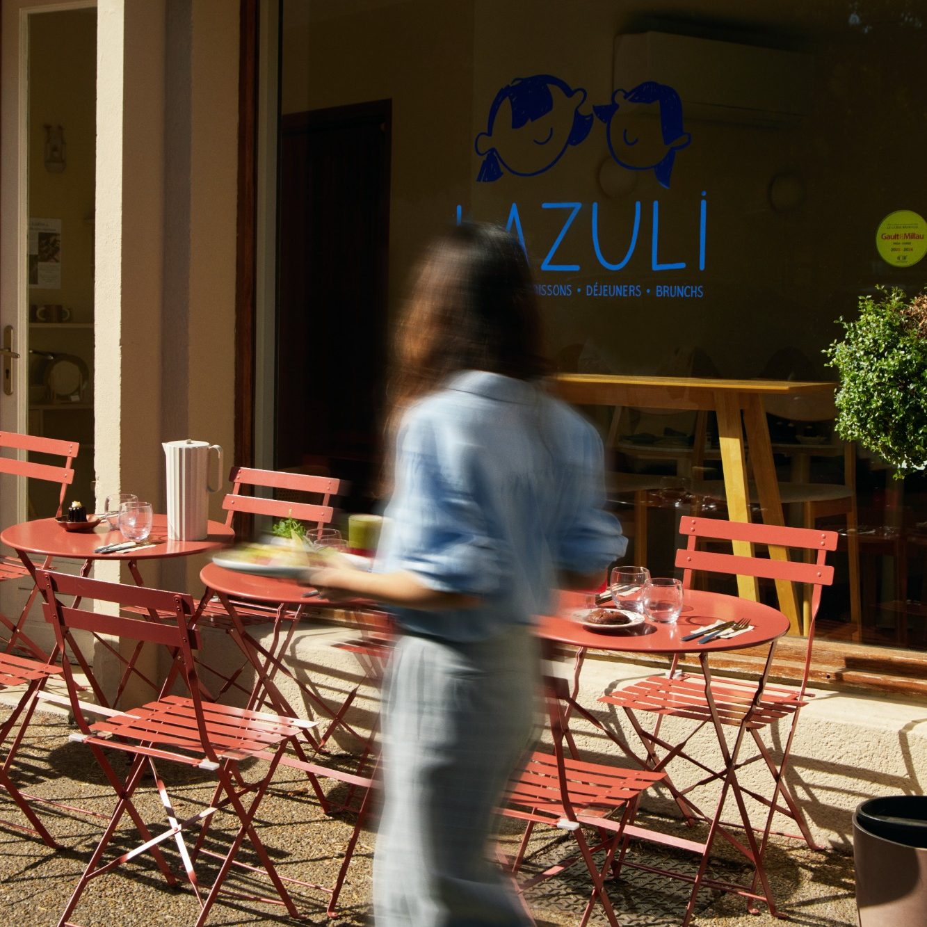 Une serveuse passe devant la vitrine LAZULI et ses petites tables terracotta en terrasse